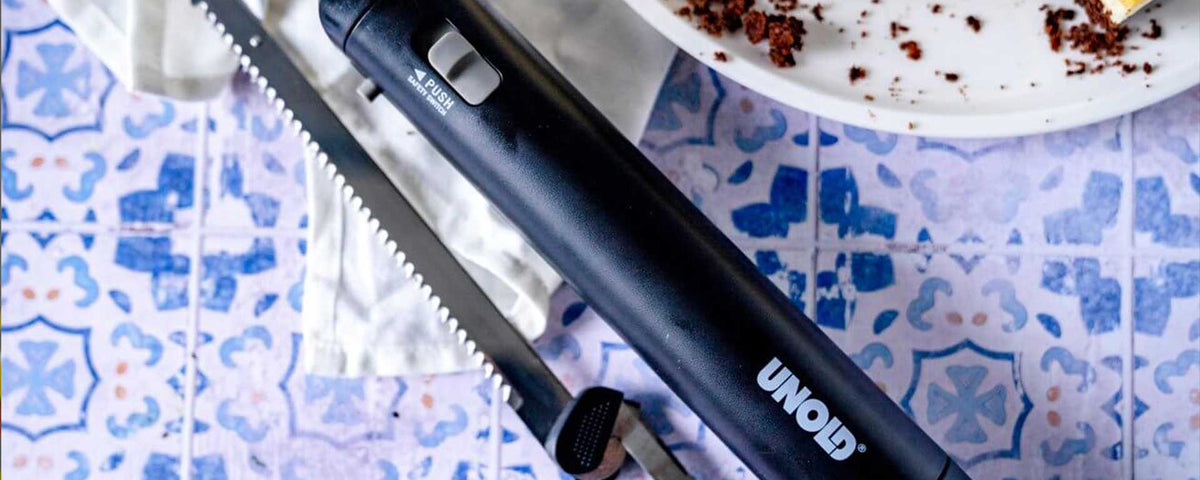 Ein schwarzes elektrisches Messer von UNOLD mit gezackter Klinge liegt auf einer gemusterten blau-weiß gefliesten Oberfläche neben einem weißen Teller mit Kuchenkrümeln.