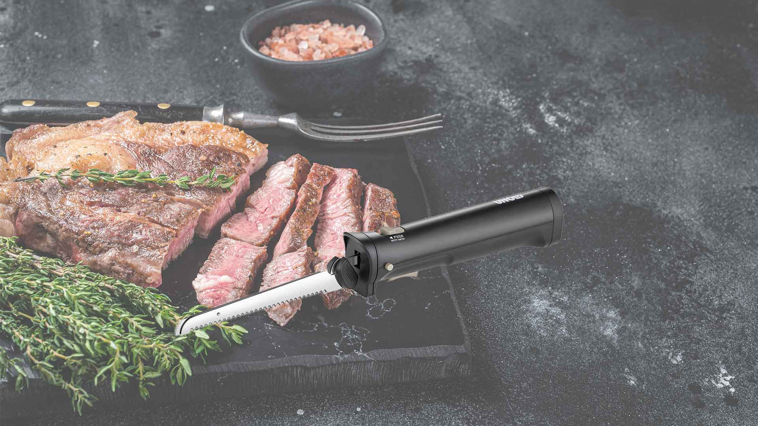 Ein mit frischen Kräutern garniertes Steak wird auf einer schwarzen Schieferplatte angerichtet, zusammen mit einer Gabel und einer Schale mit rosa Salz. Neben dem Steak liegt ein elektrisches Tranchiermesser.