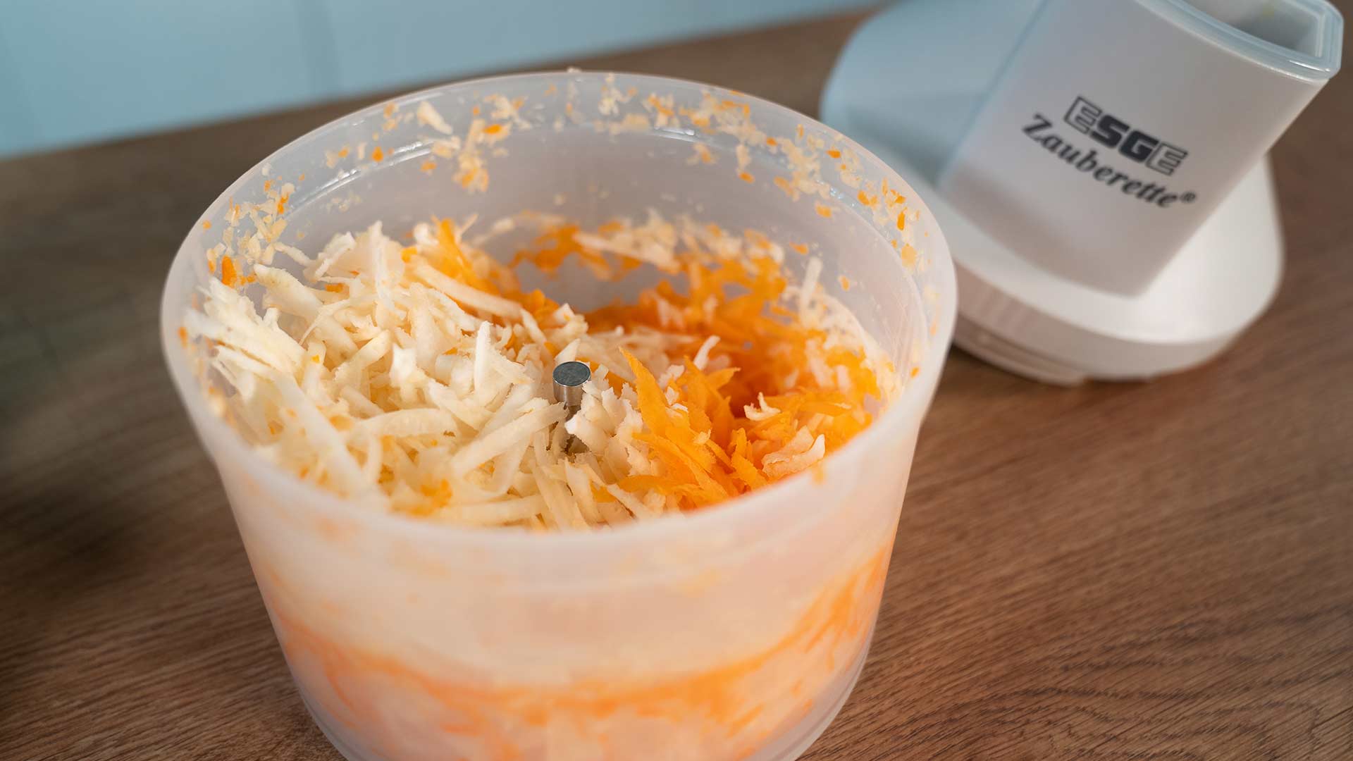 Ein Plastikbehälter mit frisch geraspeltem weißem und orangefarbenem Gemüse, wahrscheinlich Kohl und Karotten, steht auf einer Holzoberfläche neben einem weißen Küchengerät mit der Aufschrift „Zauberette“.