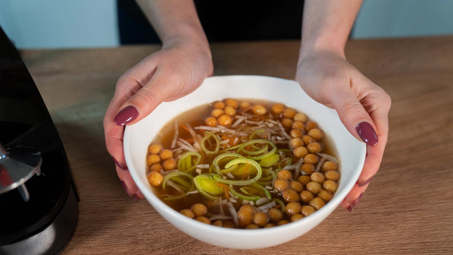 Eine Person mit lackierten Nägeln hält eine weiße Schüssel mit Suppe aus Kichererbsen, Lauchscheiben und geraspeltem Gemüse auf einem Holztisch.