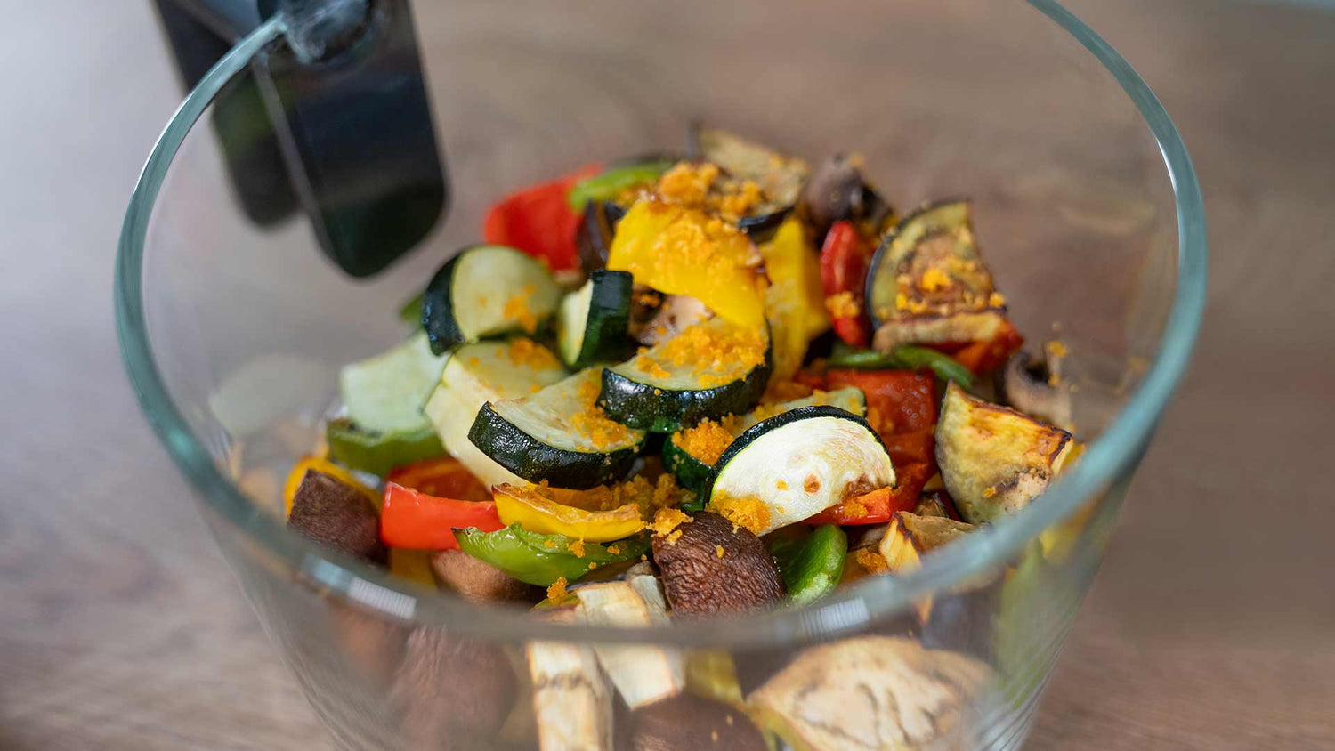 Eine Glasschale gefüllt mit gehacktem, geröstetem Gemüse, darunter Zucchini, Paprika, Pilze und Tomaten, über das eine dunkle Flüssigkeit gegossen wird.