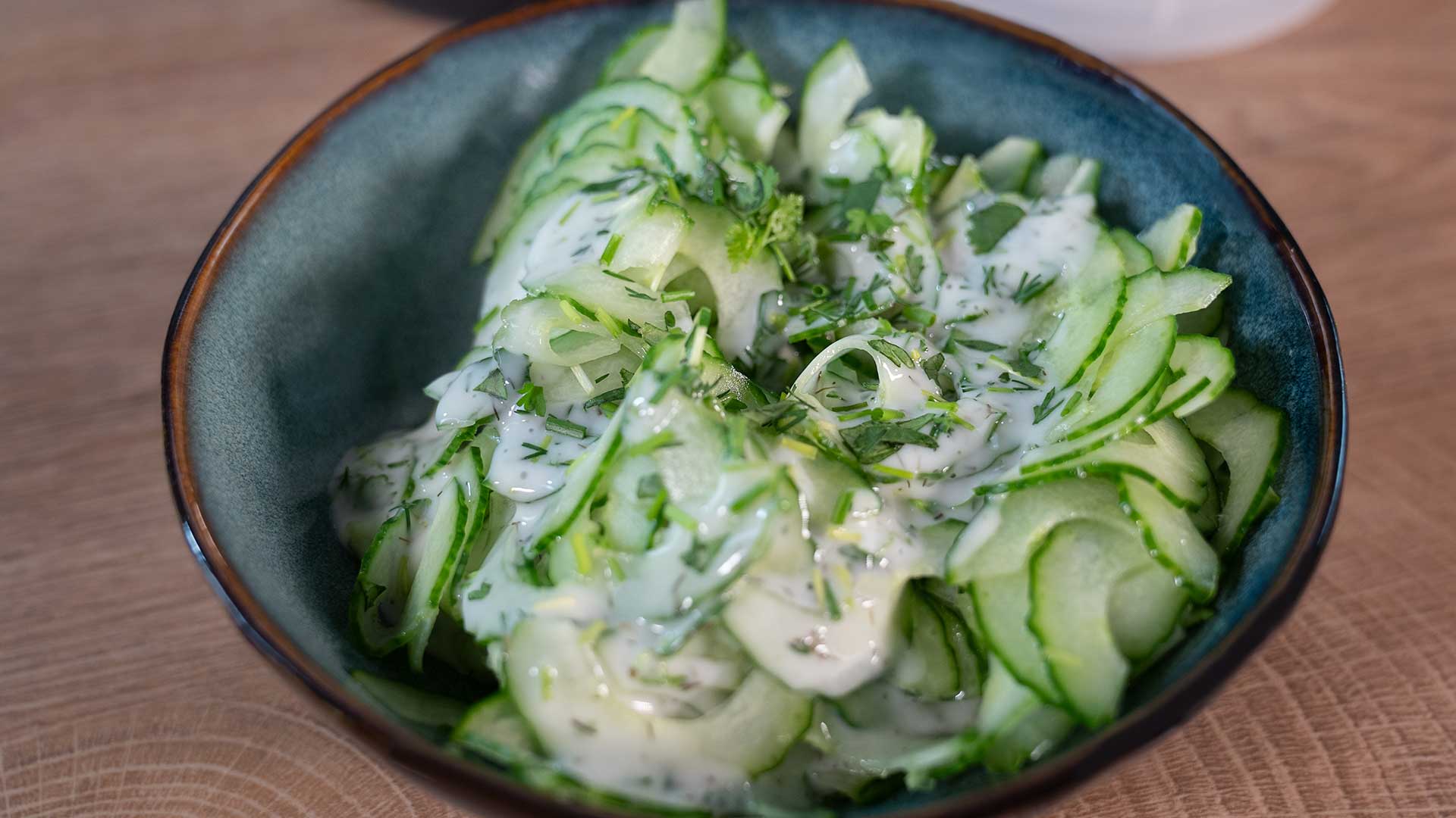 Eine Schüssel Gurkensalat mit cremigem Dressing und frischem Dill bestreut, serviert in einer Keramikschüssel mit dunklem Rand auf einer Holzoberfläche.
