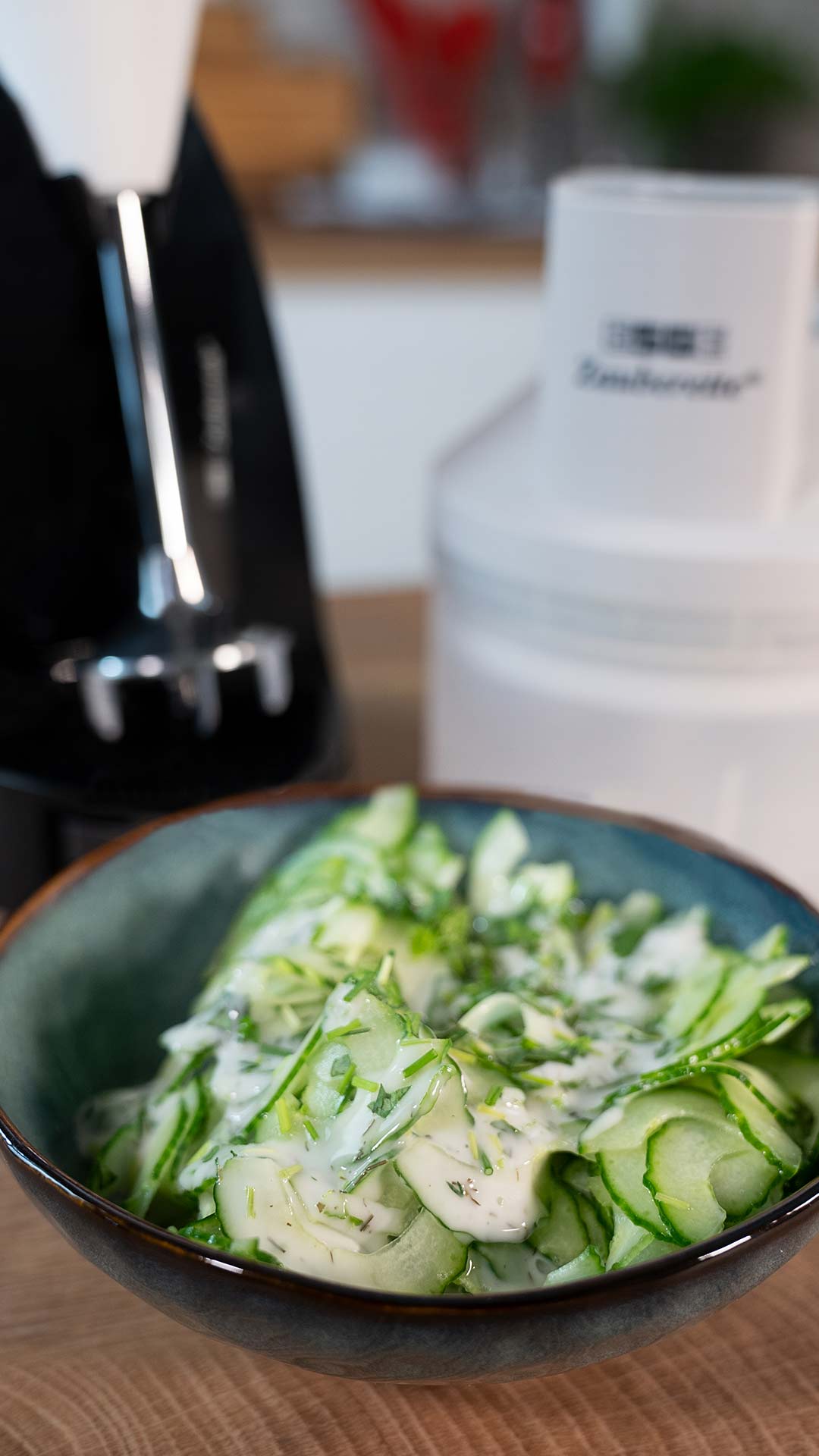 Eine Schüssel mit Gurkenscheiben und cremigem Dilldressing steht auf einer Holztheke. Im Hintergrund sind ein Mixer und sein weißer Aufsatz leicht unscharf zu sehen.