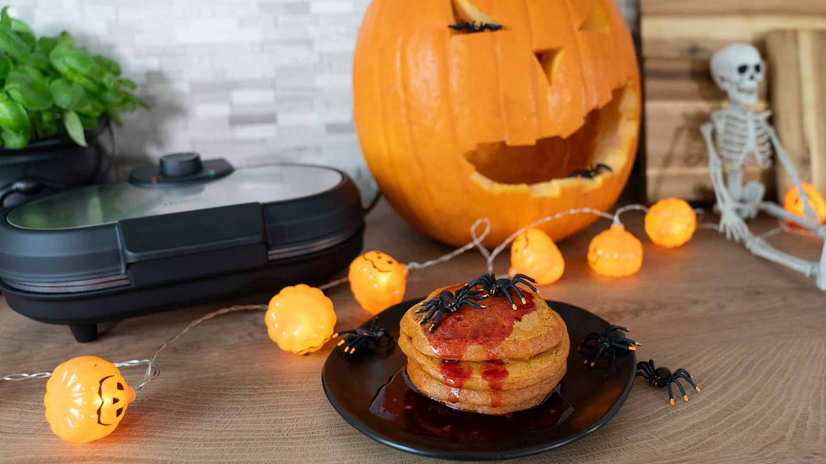 Ein Stapel Pfannkuchen mit rotem Sirup und Plastikspinnen auf einem Teller. Im Hintergrund sind eine geschnitzte Kürbislaterne, Halloween-Kürbislichter, eine Skelettdekoration und ein Küchengerät auf einer Holztheke zu sehen.