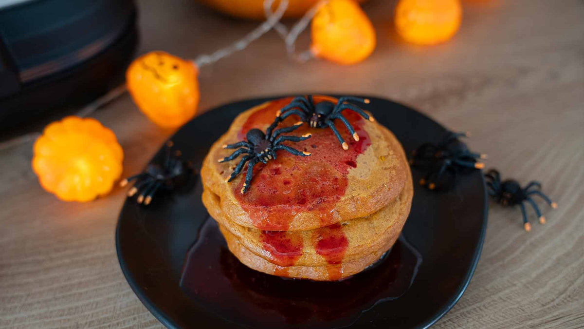 Ein Stapel Pfannkuchen mit rotem Sirup und schwarzen Plastikspinnen liegt auf einem schwarzen Teller. Im Hintergrund sorgen orangefarbene Lichter in Kürbisform für eine festliche Halloween-Atmosphäre.