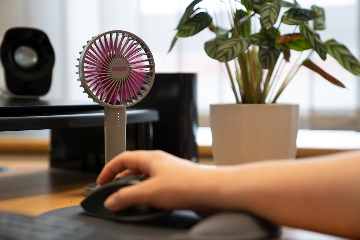 Eine Hand bedient eine Computermaus auf einem Schreibtisch neben dem Unold Handventilator Breezy II, einer Topfpflanze und Computerausrüstung, während im Hintergrund Sonnenlicht durch ein Fenster strömt.