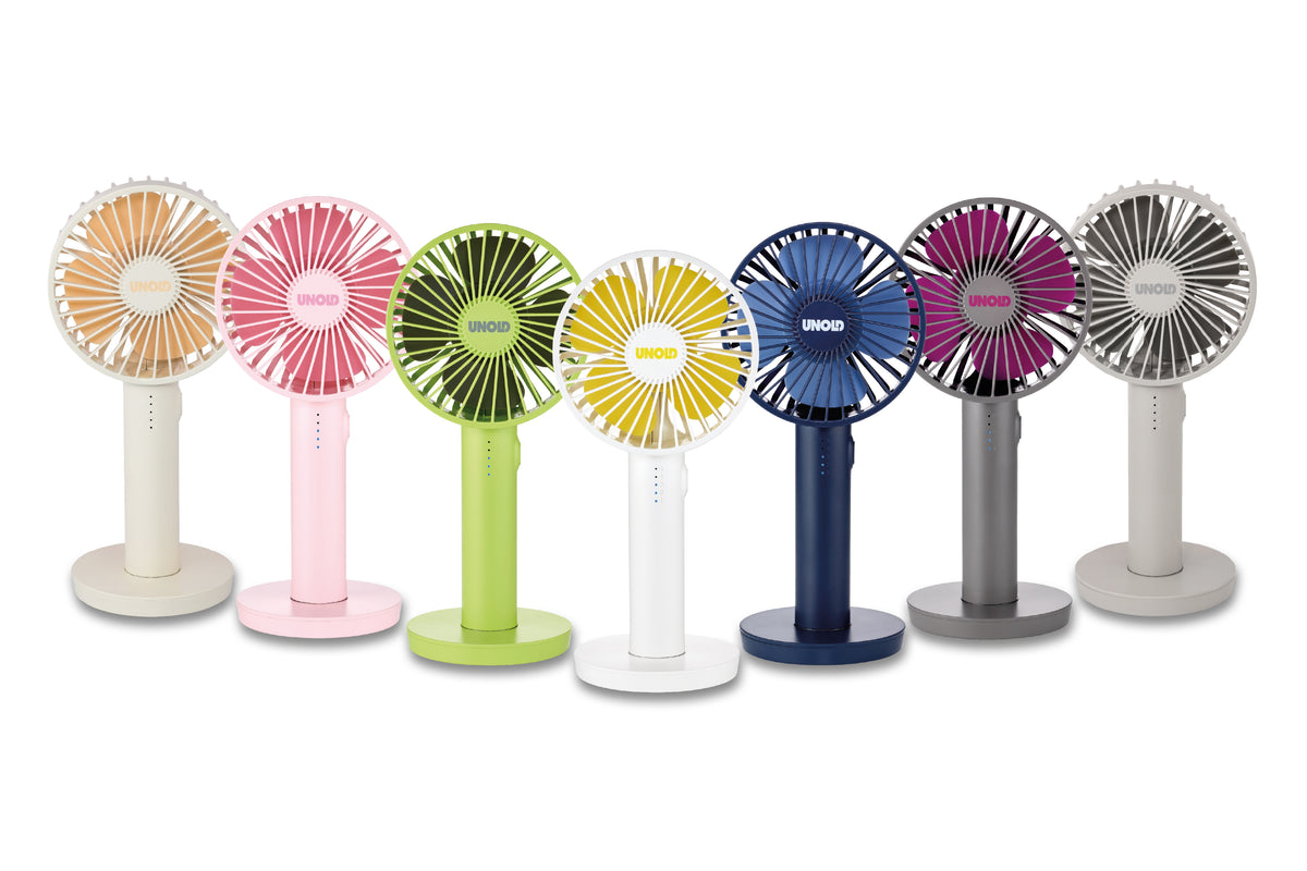 Sieben Unold Handventilator Breezy II Ventilatoren in den Farben Pfirsich, Rosa, Grün, Gelb, Weiß, Blau, Lila und Grau sind auf einem weißen Hintergrund aufgereiht. Jeder tragbare Handventilator hat einen runden Kopf mit einem radialen Flügeldesign.