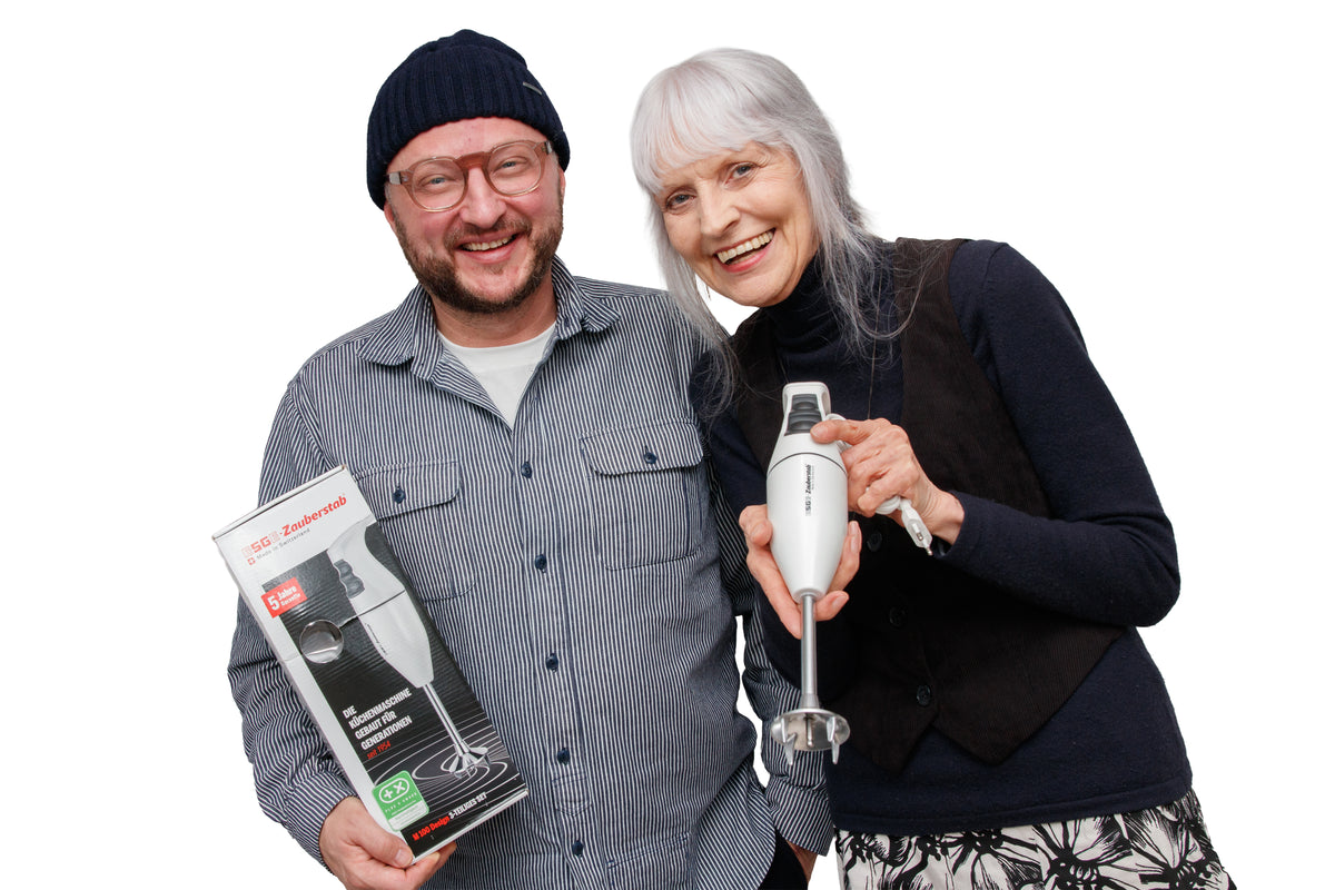 Ein lächelnder Mann mit einer Mütze hält eine Schachtel für einen Stabmixer, während eine lächelnde ältere Frau mit langen grauen Haaren den Mixer selbst hält. Beide stehen dicht beieinander vor einem weißen Hintergrund.