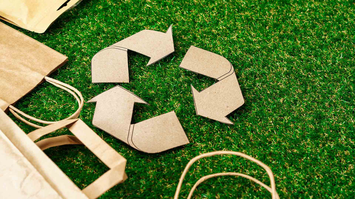 Auf grünem Gras liegt ein Recyclingsymbol aus Pappe, umgeben von braunem Papier und Kordelgriffen, was auf umweltfreundliche und nachhaltige Praktiken hindeutet.