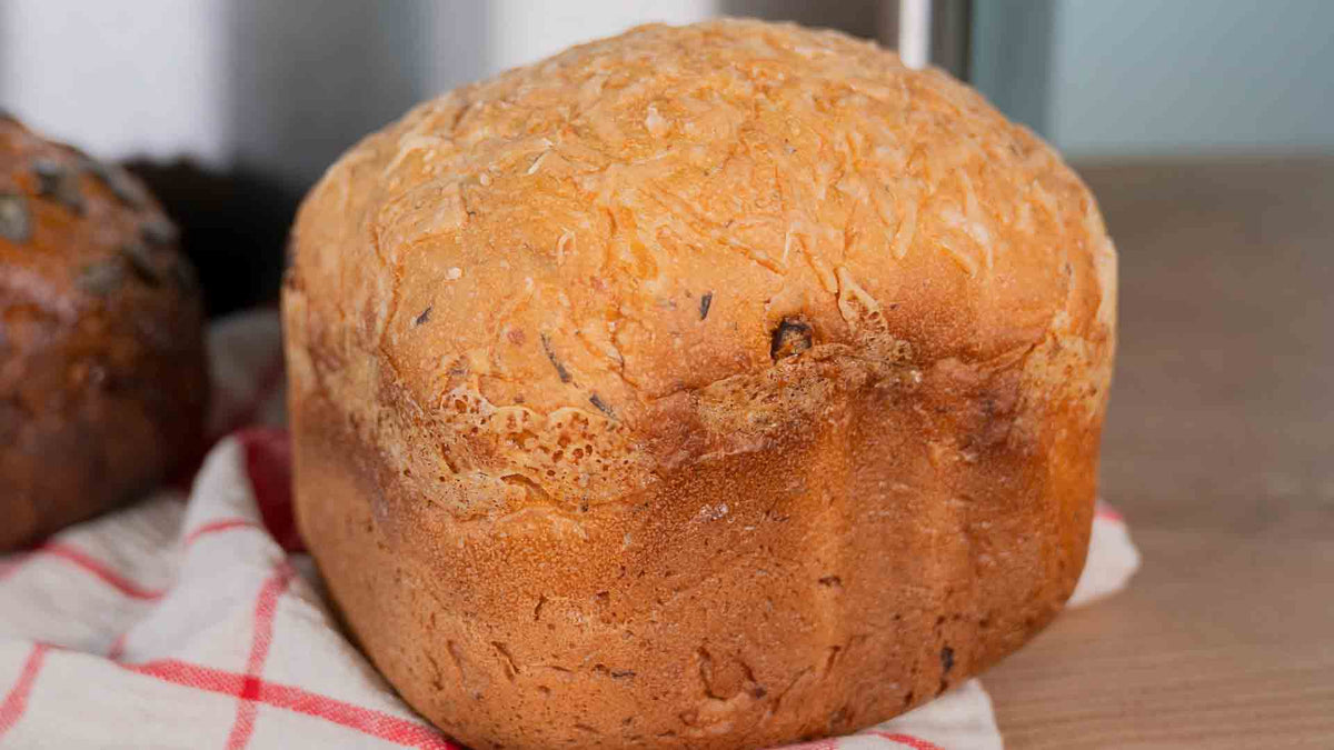 Ein goldbrauner Laib Brot mit strukturierter Oberseite liegt auf einem rot-weiß karierten Tuch auf einer Holzoberfläche.