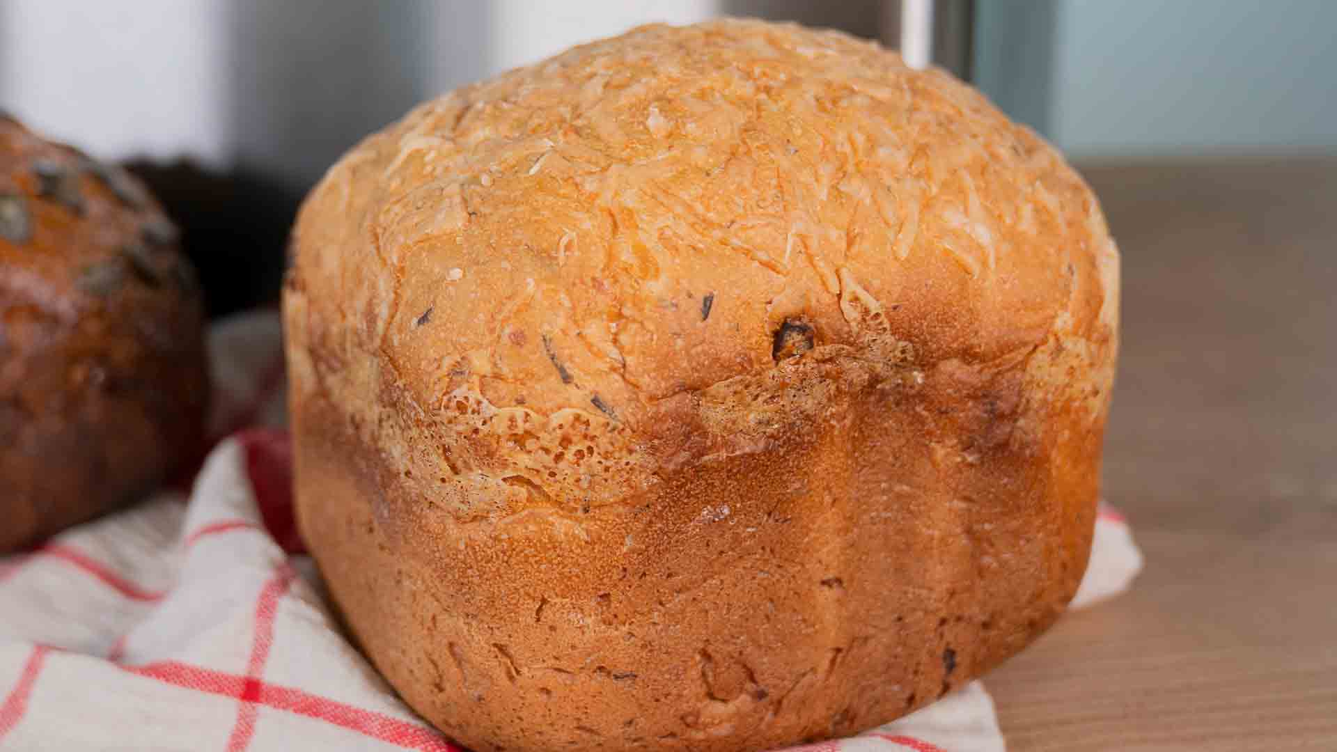 Ein goldbrauner Laib Brot mit strukturierter Oberseite liegt auf einem rot-weiß karierten Tuch auf einer Holzoberfläche.