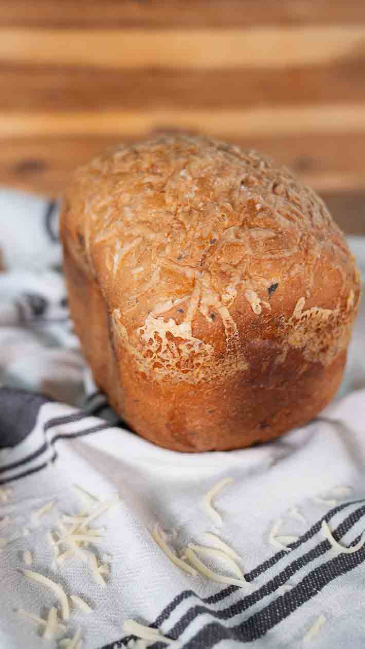 Ein goldbrauner Laib Brot mit geschmolzenem Käse liegt auf einem weißen Handtuch mit schwarzen Streifen, darüber sind einige Käsesplitter verstreut. Der Hintergrund ist leicht verschwommenes Holz.