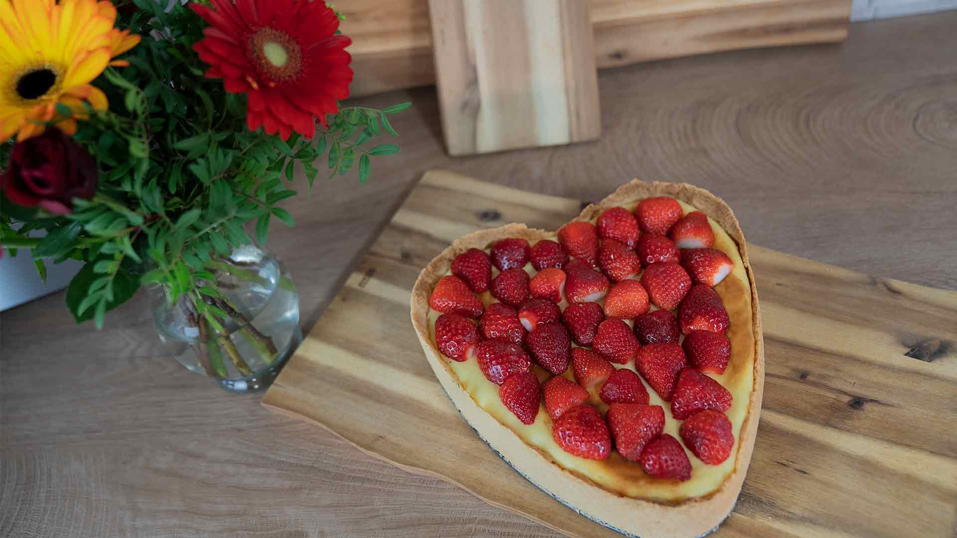 Ein herzförmiger Käsekuchen mit frischen Erdbeeren steht auf einem Holzschneidebrett neben einer Vase mit bunten Blumen auf einer Holzarbeitsplatte.