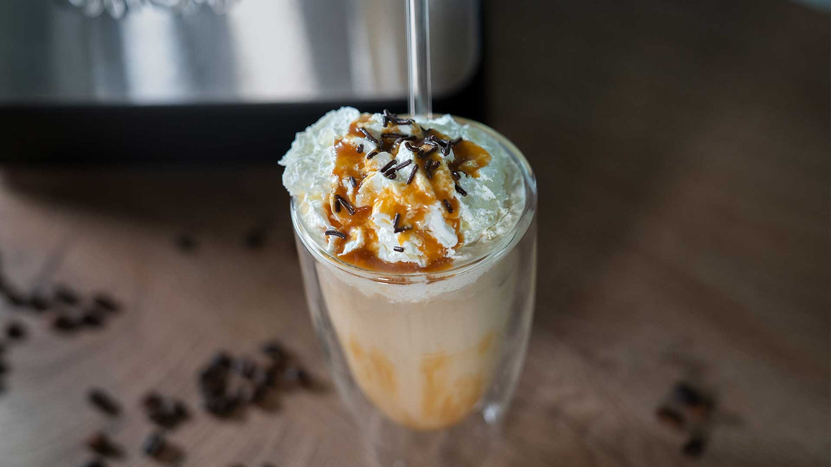 Ein Glas Eiskaffee mit Schlagsahne, Karamellsauce und Schokoladenraspeln sowie einem Strohhalm steht auf einer Holzoberfläche, auf der Kaffeebohnen verstreut sind.