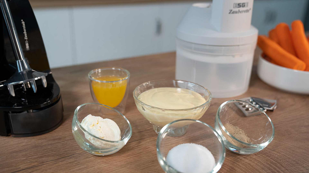 Eine Holzarbeitsplatte mit kleinen Glasschüsseln, die weiße und cremige Zutaten, Orangensaft und Gewürze enthalten. Daneben stehen ein Stabmixer, eine Küchenmaschine, ein Gemüseschäler und eine Schüssel mit ganzen Karotten.