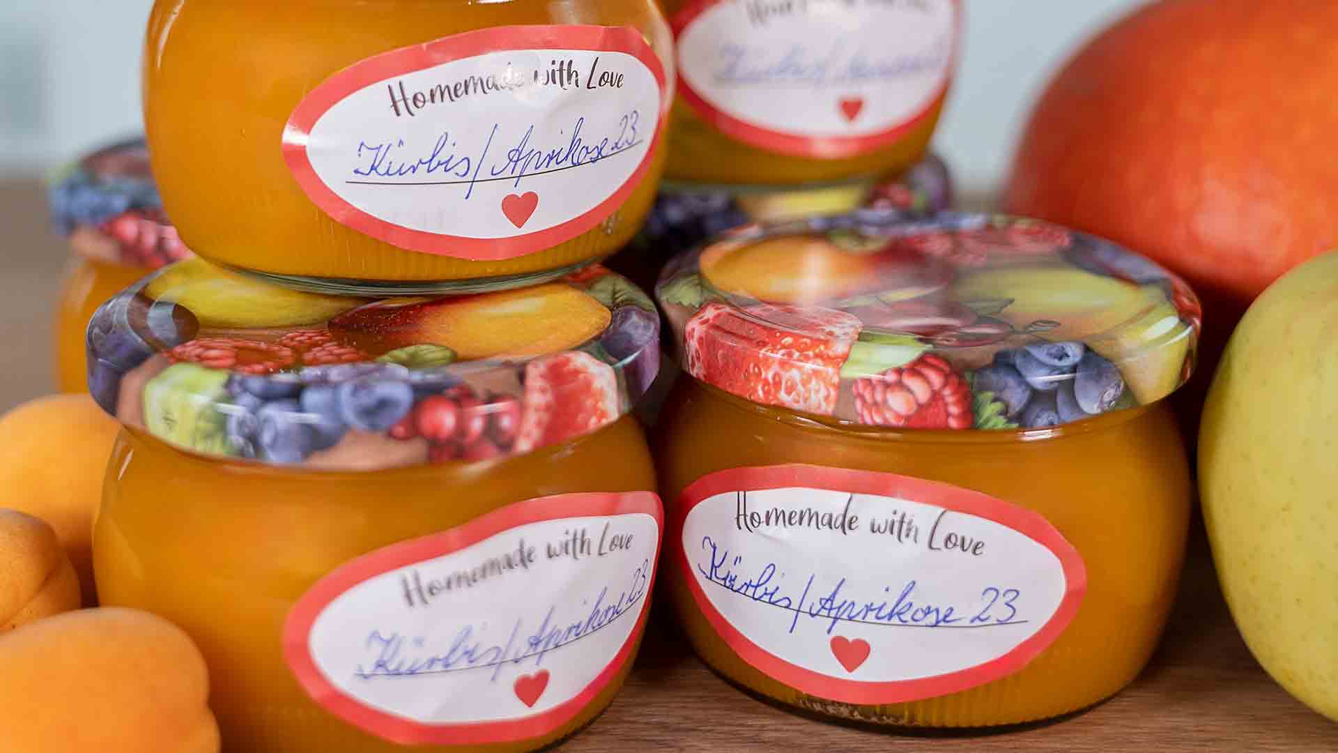 Mehrere Gläser mit selbstgemachter Marmelade, bunten Deckeln mit Fruchtmustern und handgeschriebenen Etiketten mit der Aufschrift „Homemade with Love Kürbis/Aprikose 23“ stehen neben frischem Obst gestapelt.