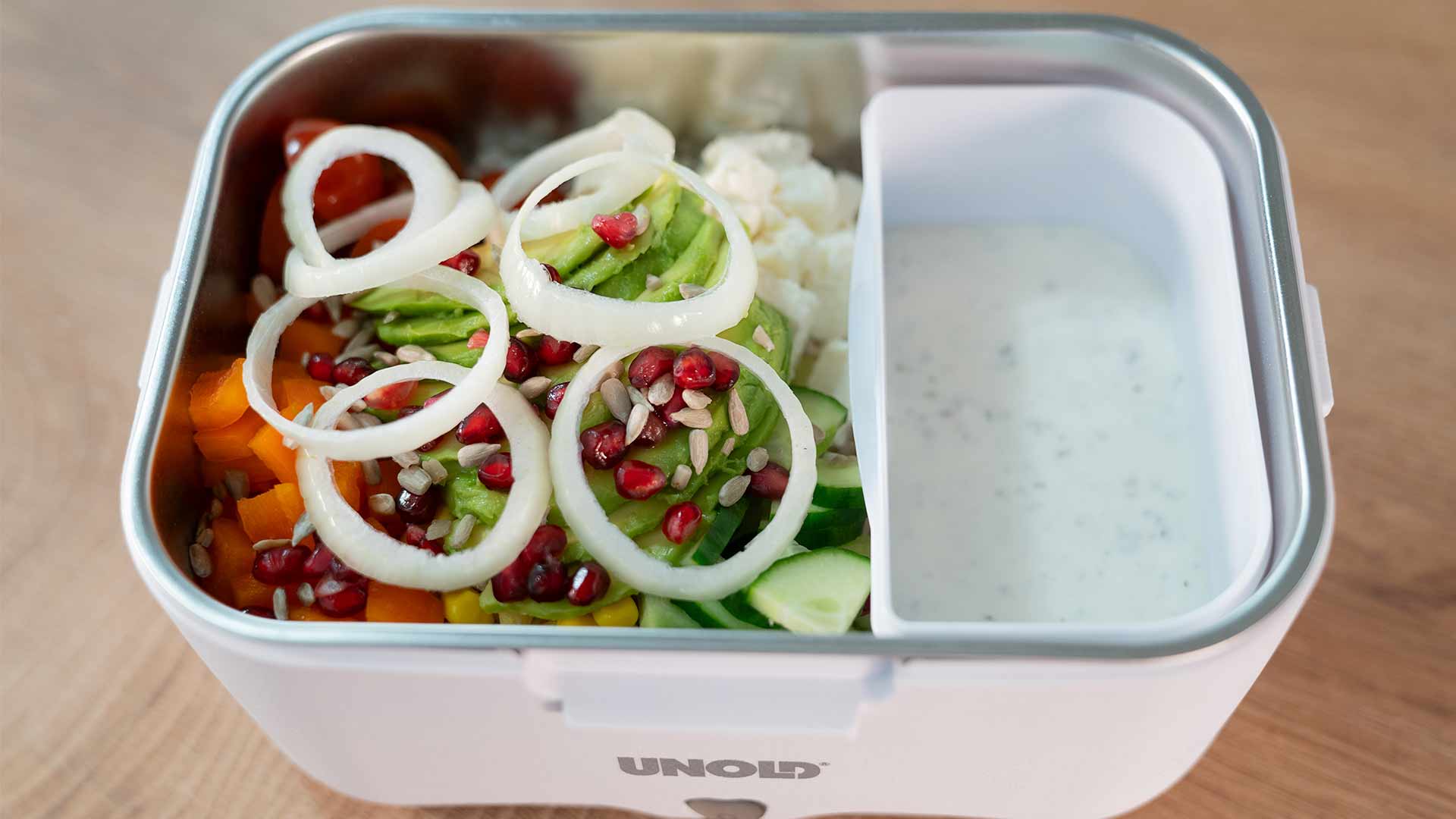 Eine Lunchbox mit einem bunten Salat aus geschnittenem Gemüse, Zwiebelringen und Granatapfelkernen auf der einen Seite und einem Fach mit cremeweißem Dressing auf der anderen Seite, platziert auf einer Holzoberfläche.