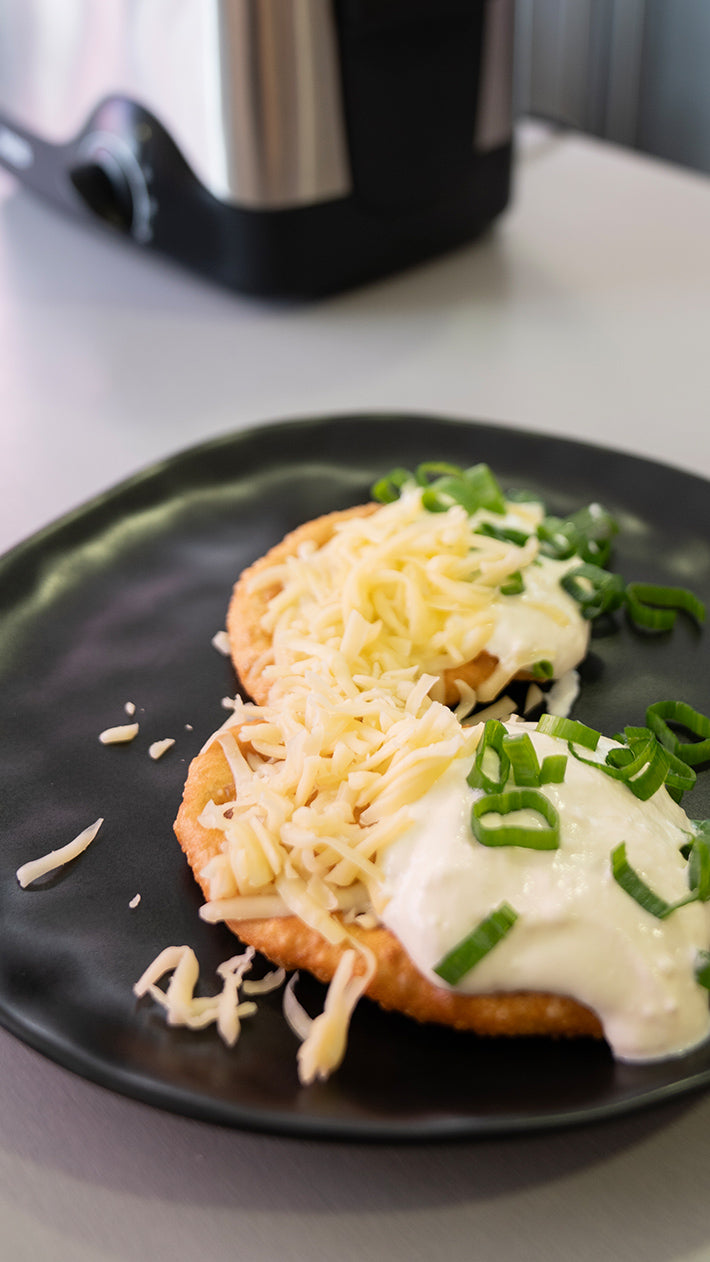 Zwei Scheiben frittiertes Brot mit Sauerrahm, geriebenem Käse und gehackten Frühlingszwiebeln werden auf einem schwarzen Teller serviert. Im Hintergrund ist ein Küchengerät unscharf zu sehen.