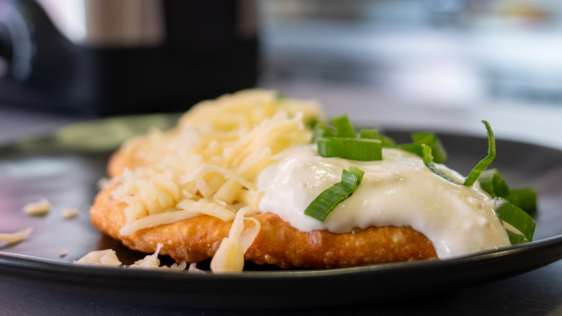 Eine Nahaufnahme eines panierten, frittierten Pastetchens mit geriebenem Käse und weißer Soße, garniert mit gehackten Frühlingszwiebeln, serviert auf einem dunklen Teller.