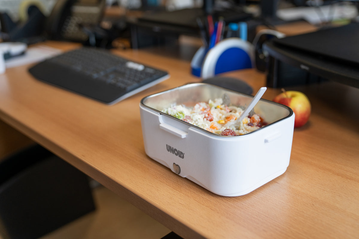 Eine Unold Lunchbox in Weiß, gefüllt mit Essen und einem Löffel, steht auf einem Bürotisch neben einer Tastatur, einer Maus und einem Apfel im Hintergrund.