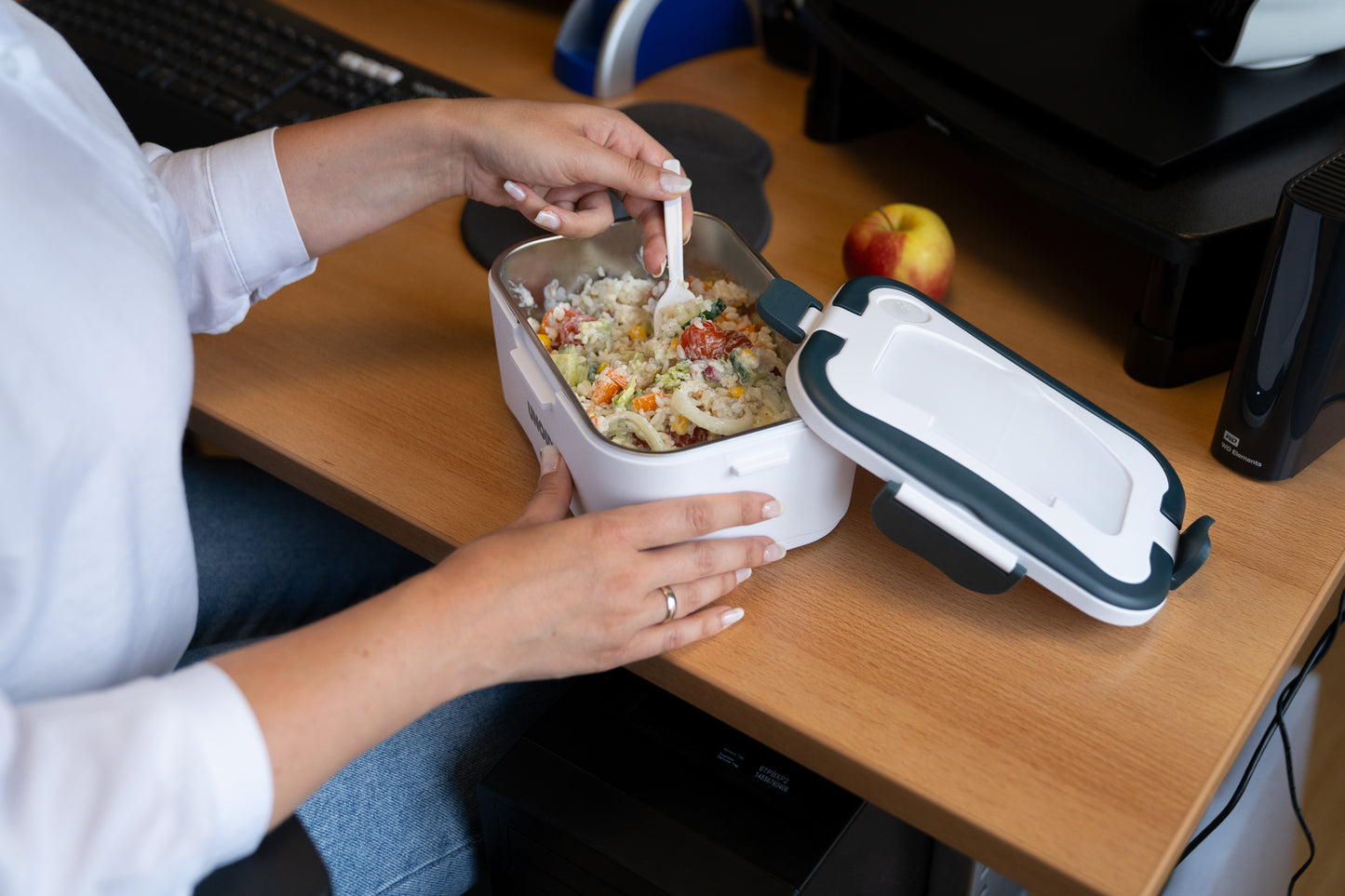 Eine Person am Schreibtisch isst mit einer Gabel Reis und Gemüse aus der Unold Lunchbox; daneben liegen der geschlossene Deckel und ein Apfel.