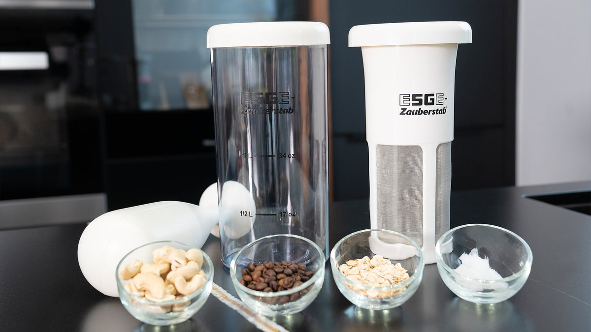 Drei Glasschüsseln mit Cashewnüssen, Kaffeebohnen und Hafer stehen vor zwei ESGE Zauberstab-Behältern, einer davon mit Metallgittereinsatz, auf einer schwarzen Küchentheke, links davon ein Stabmixer.