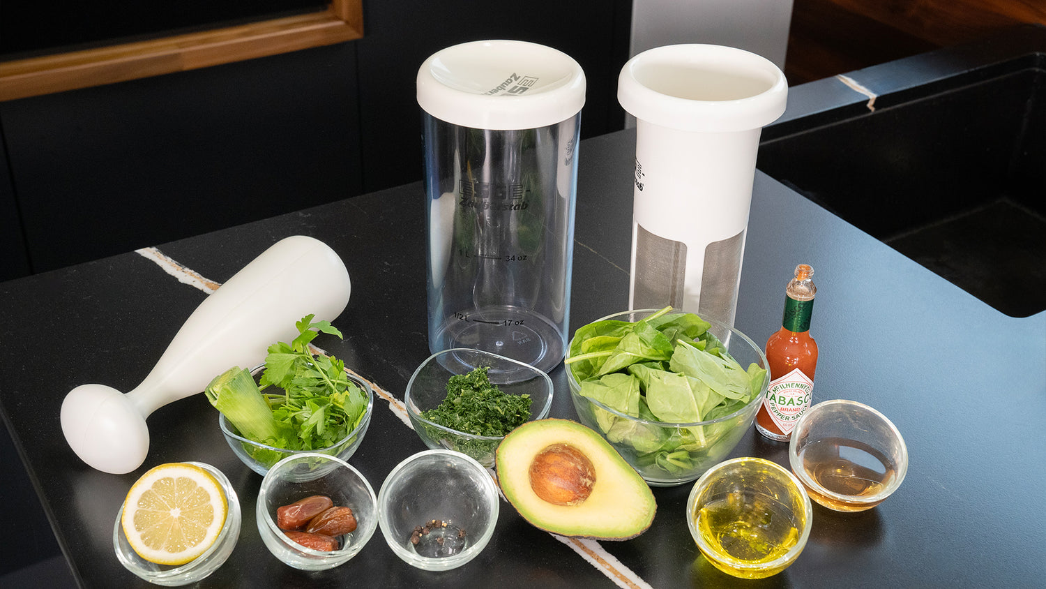 Verschiedene frische Zutaten, darunter eine halbe Avocado, eine Zitronenscheibe, Spinat, Petersilie, Mandeln, Öl und Gewürze, sind auf einer schwarzen Arbeitsplatte mit einem Stabmixer und Mixbehältern angeordnet.