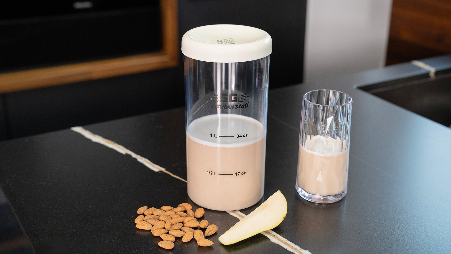 Eine Szene auf einer Arbeitsplatte mit einem durchsichtigen Mixbecher, der einen beigen Smoothie enthält, einem Glas mit noch mehr Smoothie, einem kleinen Haufen roher Mandeln und einer Birnenspalte, alles angeordnet auf einer schwarzen Marmoroberfläche.