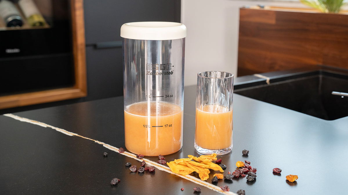 Ein hoher Glaskrug und ein Glas, beide mit Orangensaft gefüllt, stehen auf einer schwarzen Arbeitsplatte. In der Nähe liegen verstreut getrocknete Fruchtstücke, im Hintergrund sind Küchenschränke und eine Spüle zu sehen.