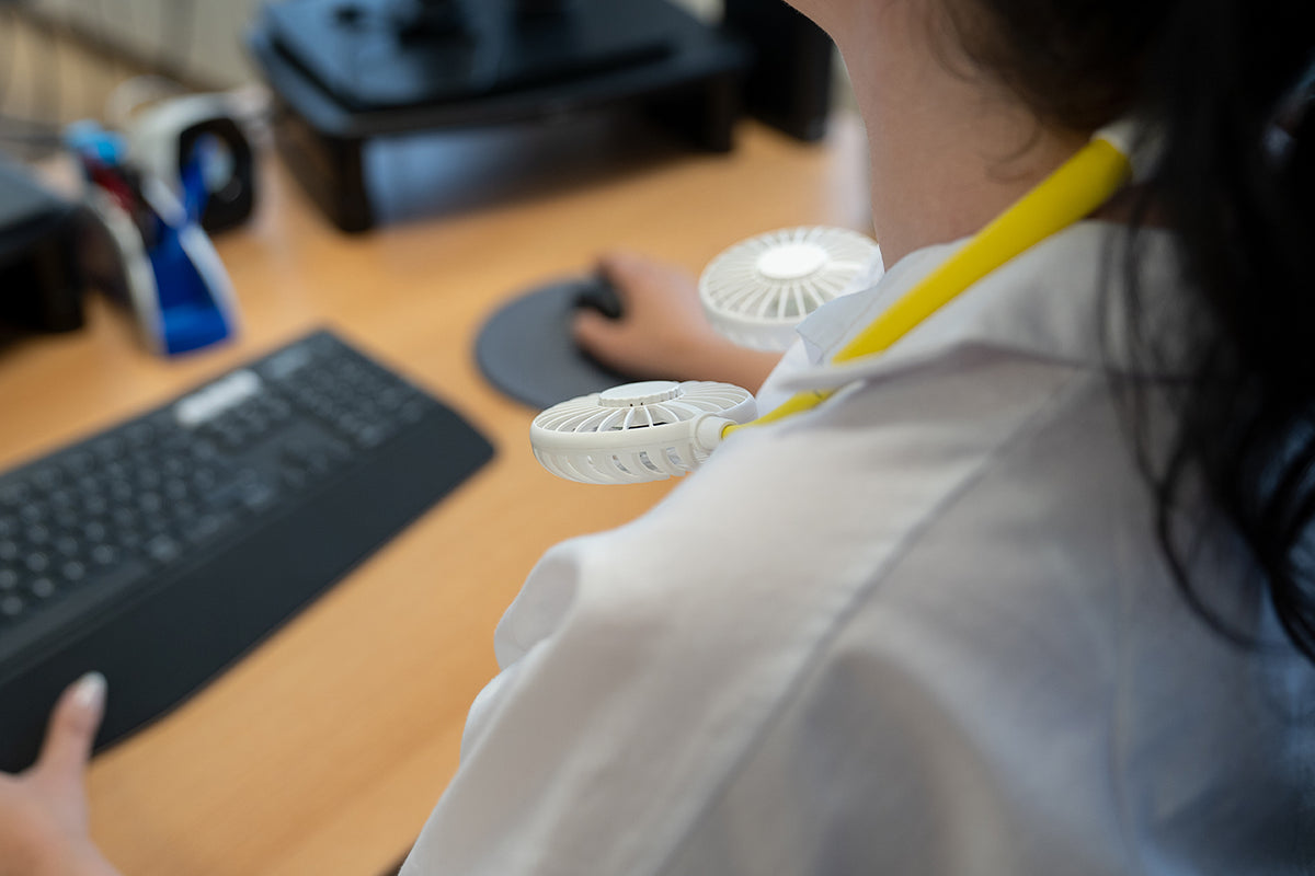 Eine Person im weißen Kittel bedient eine Computermaus mit dem Unold NACKENVENTILATOR BREEZY, einem tragbaren flügellosen Nackenventilator, um den Hals; Tastatur und Monitor stehen auf dem Holzschreibtisch.