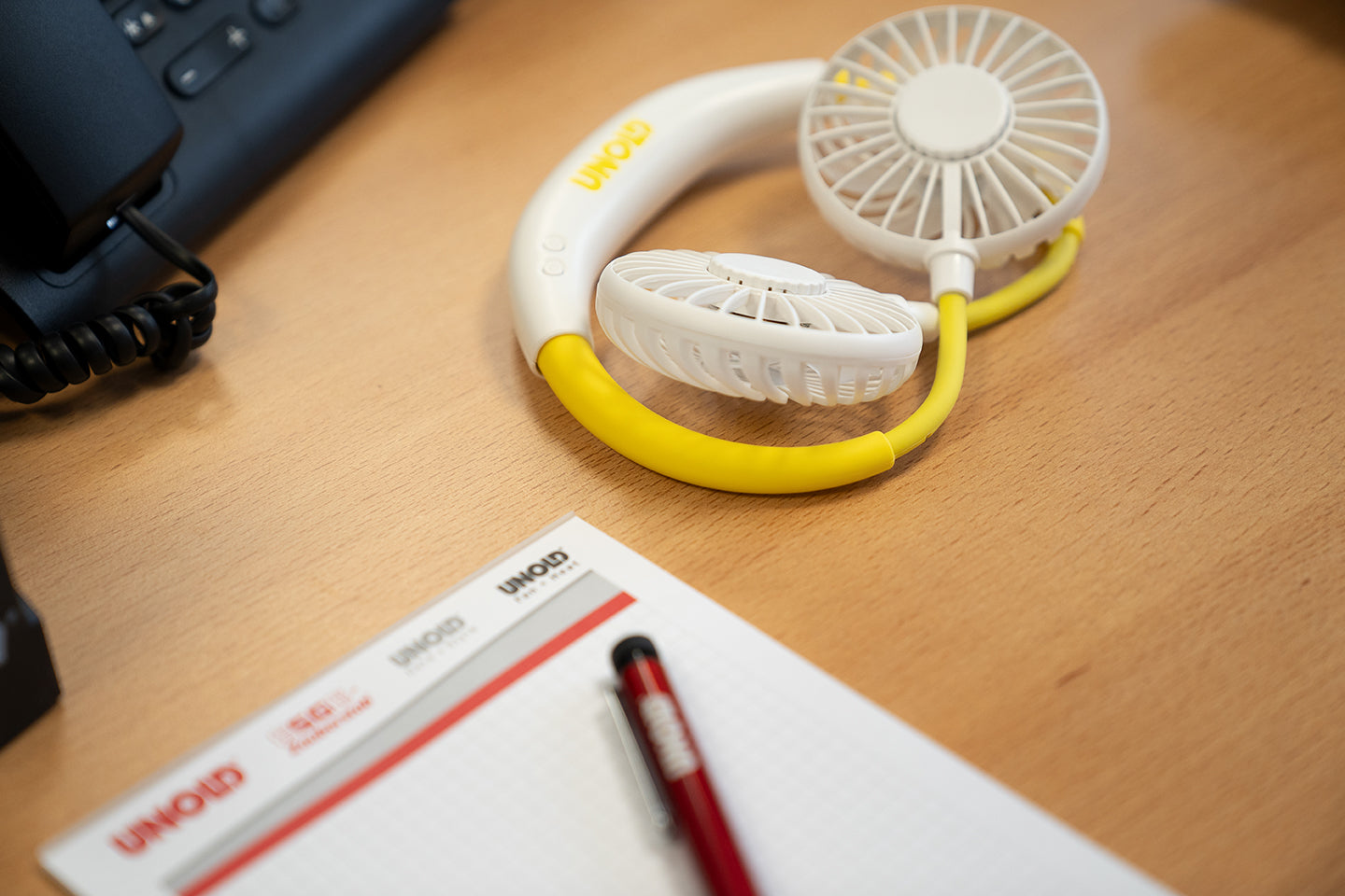 Ein Nackenventilator NACKENVENTILATOR BREEZY von Unold mit gelb-weißen Akzenten steht neben einem UNOLD-Notizblock und einem roten Stift auf einem Holzschreibtisch. Links ist ein Teil eines schwarzen Bürotelefons zu sehen.
