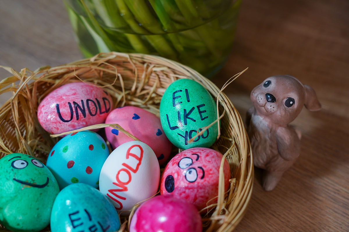 Ein Korb mit bunten, verzierten Ostereiern mit Botschaften wie UNOLD und EI LIKE YOU, daneben eine kleine, braune Hundefigur aus Keramik. Im Hintergrund steht eine Vase mit grünen Stielen.