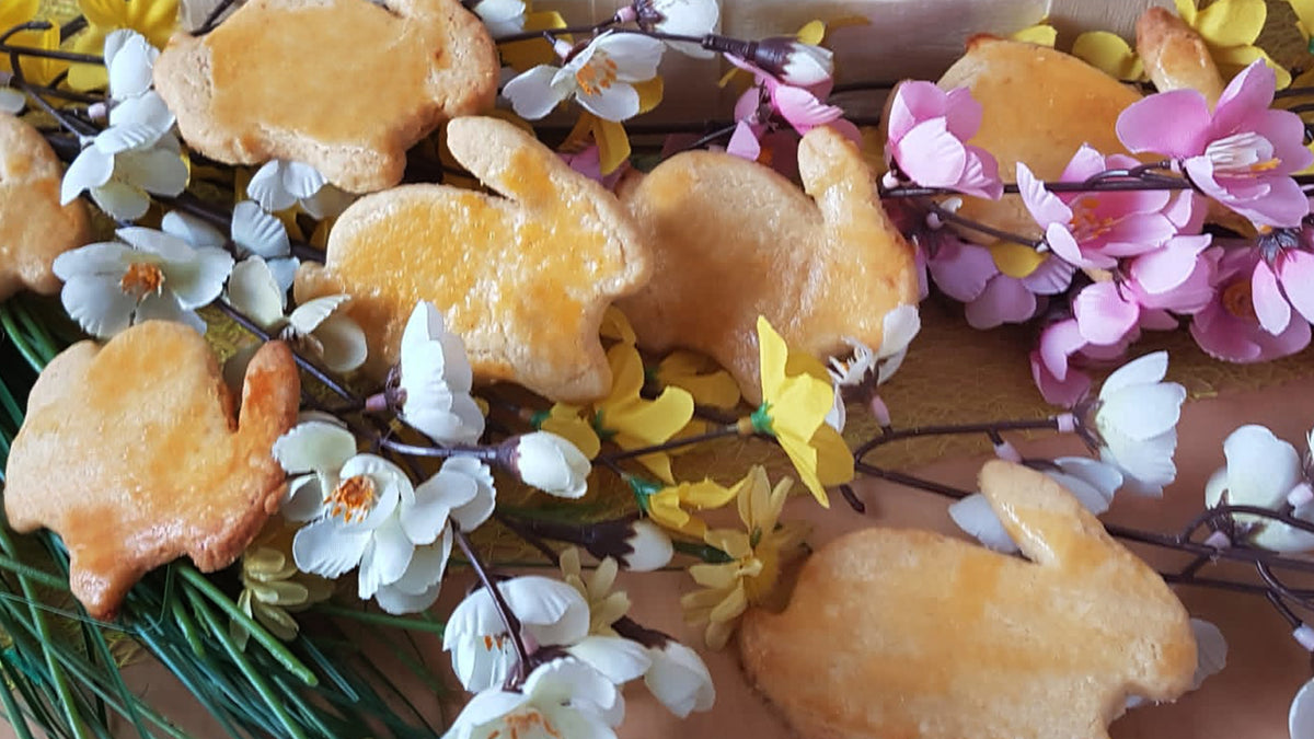 Kekse in Form eines Osterhasen, angeordnet zwischen künstlichen rosa, gelben und weißen Blumen und grünen, grasähnlichen Stängeln auf einer Holzoberfläche.