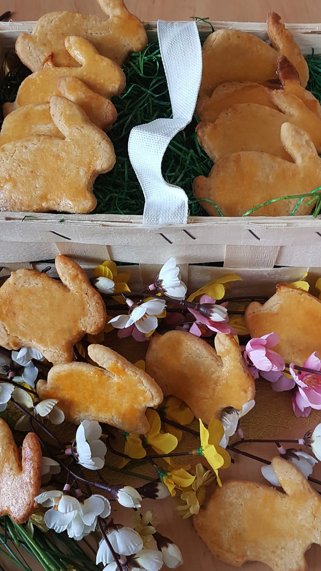 Ein Holzkorb gefüllt mit hasenförmigen Keksen, einige davon auf Kunstrasen und andere zwischen pastellfarbenen und weißen Kunstblumen auf einer Holzoberfläche angeordnet.