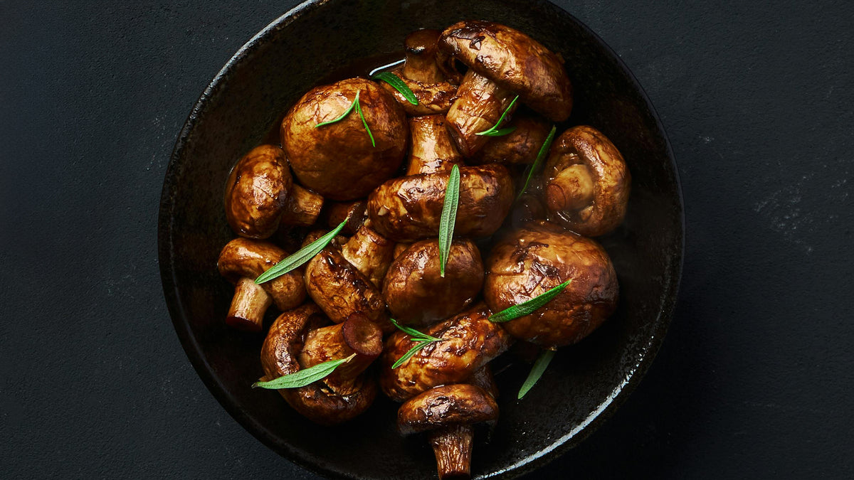 Eine Schüssel gefüllt mit gekochten braunen Champignons, garniert mit frischen grünen Kräutern, vor einem dunklen Hintergrund.