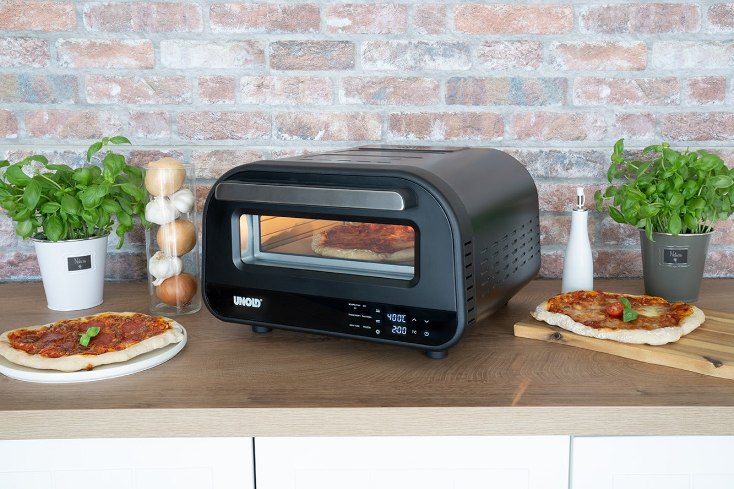 Der Unold B-Ware Pizzaofen Don Luigi in Schwarz steht auf einer Holzfläche neben zwei Basilikumpflanzen, Zwiebeln, Salz- und Pfefferstreuern und zwei fertigen Pizzen auf Schneidebrettern, im Hintergrund eine Backsteinmauer.
