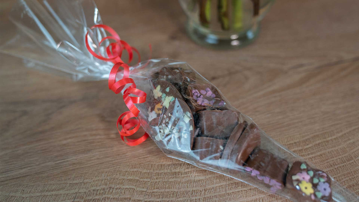 Ein durchsichtiger Plastikbeutel, gefüllt mit Schokoladenstückchen und mit bunten Mustern verziert, wird mit einem roten Schnörkelband zugebunden und auf eine Holzoberfläche gelegt.