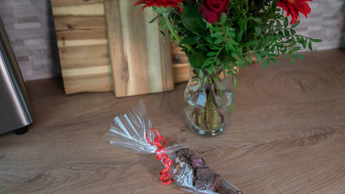 Ein Strauß roter Blumen in einer Glasvase steht auf einer Holztheke neben einer durchsichtigen Tüte mit verschiedenen Pralinen, die mit einer roten Schleife zusammengebunden ist. Im Hintergrund sind hölzerne Schneidebretter zu sehen.