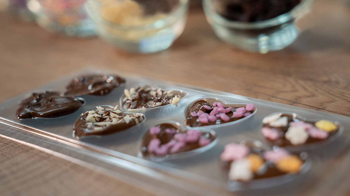 Eine Nahaufnahme einer durchsichtigen Form mit herzförmigen Pralinen, verziert mit bunten Streuseln und Schokoladenraspeln, liegt auf einer Holzoberfläche. Im Hintergrund sind verschwommene Schüsseln mit Toppings zu sehen.
