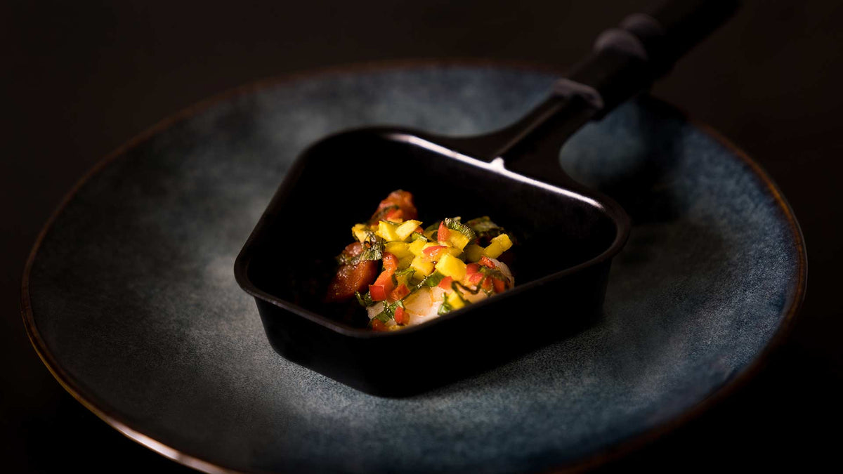 Auf einem dunkelblauen Teller liegt ein schwarzer Löffel, auf dem sich eine kleine Portion fein gewürfeltes Gemüse und Kräuter befindet, kunstvoll angerichtet als Gourmet-Vorspeise.