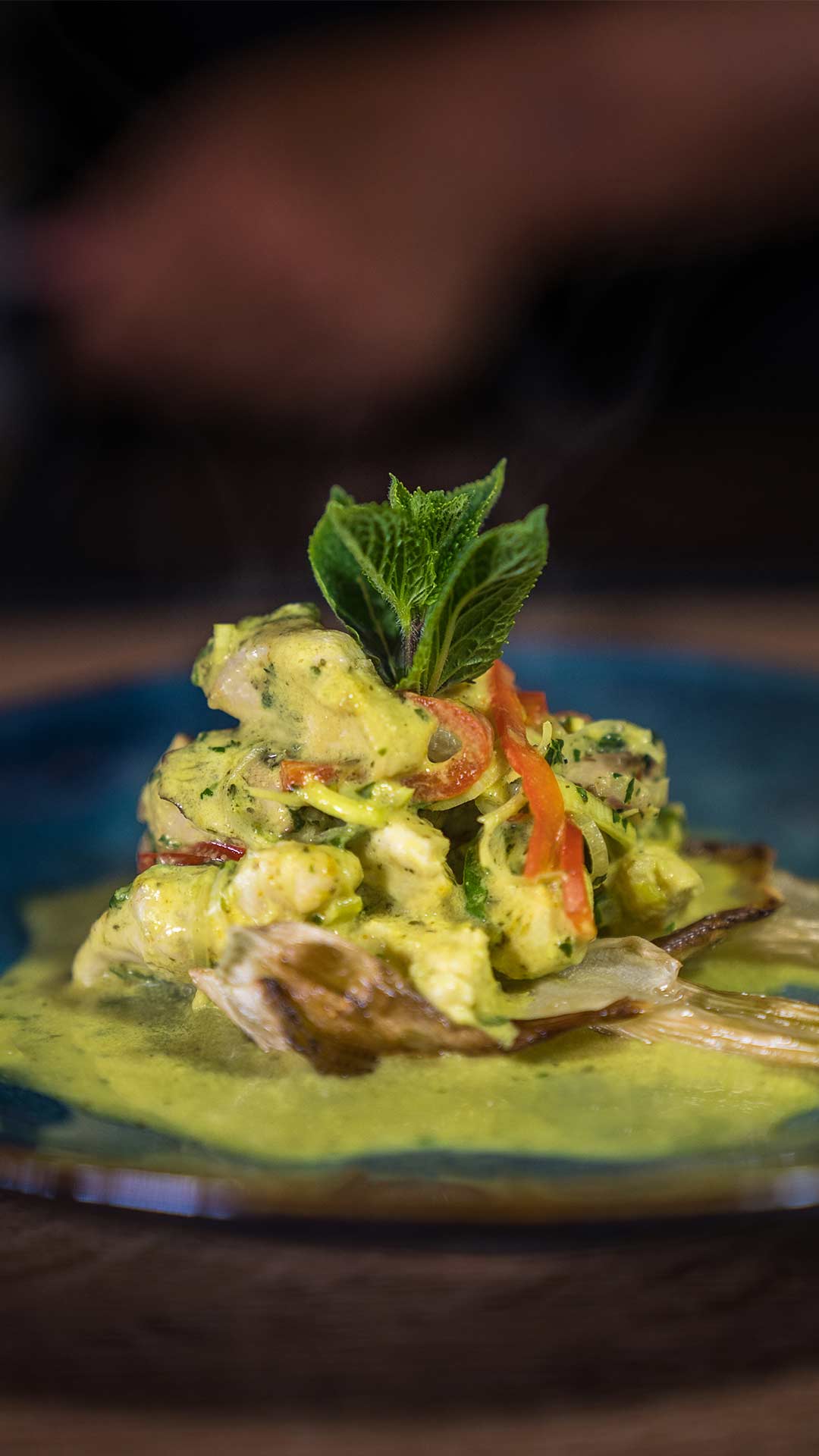 Eine Nahaufnahme eines Gourmetgerichts mit cremigen, mit Kräutern gewürzten Meeresfrüchten oder Hühnchen, garniert mit roten Paprika und frischen Minzblättern, serviert auf einem blauen Teller mit grüner Soße.