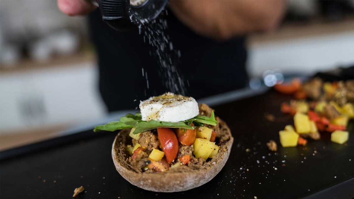 Eine Hand streut Pfeffer über einen gefüllten Champignonhut mit gewürfeltem Gemüse und Rucola, garniert mit einer Scheibe Käse auf einer Küchentheke.