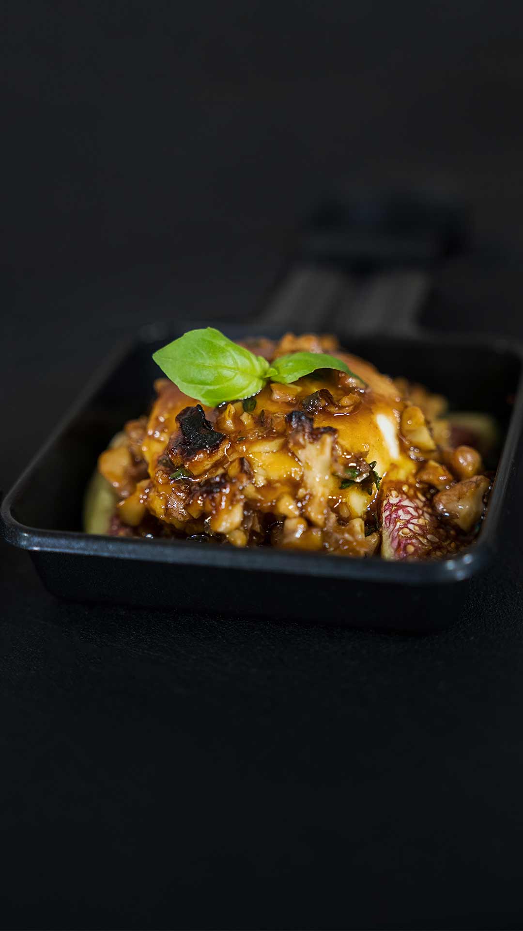 Eine Nahaufnahme eines Gourmetgerichts in einer schwarzen quadratischen Pfanne, garniert mit einer reichhaltigen Sauce, gehackten Nüssen oder Gemüse und einem kleinen frischen Basilikumblatt vor einem dunklen Hintergrund.