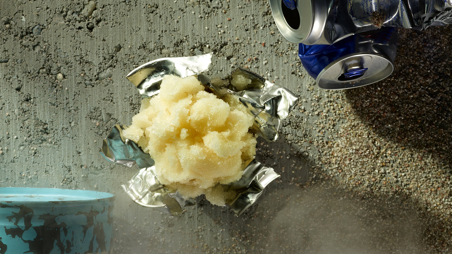 Eine zerdrückte Aluminiumdose mit gelblichem Eis oder Matsch im Inneren liegt auf einem rauen, sandigen Untergrund. Eine weitere zerdrückte blaue Dose und ein blaues Fass stehen in der Nähe.