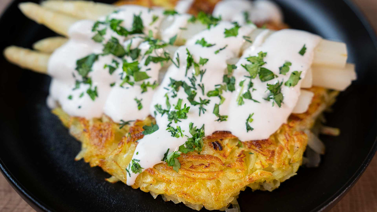 Ein Teller mit goldbraunem, knusprigem Rösti, belegt mit weißen Spargelstangen, beträufelt mit cremiger weißer Sauce und garniert mit gehackter frischer Petersilie.