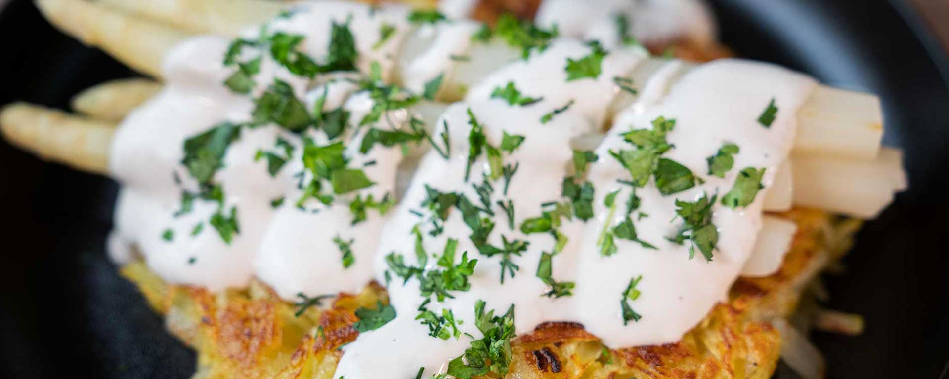 Eine Nahaufnahme von weißem Spargel mit cremiger weißer Sauce und gehackten frischen Kräutern, serviert auf einer goldbraunen Kartoffelrösti.