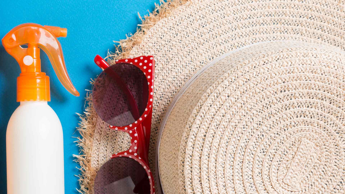 Eine Nahaufnahme eines geflochtenen Strohhuts, einer rot gepunkteten Sonnenbrille und einer weißen Sprühflasche mit orangefarbener Düse, angeordnet auf einem blauen Hintergrund.