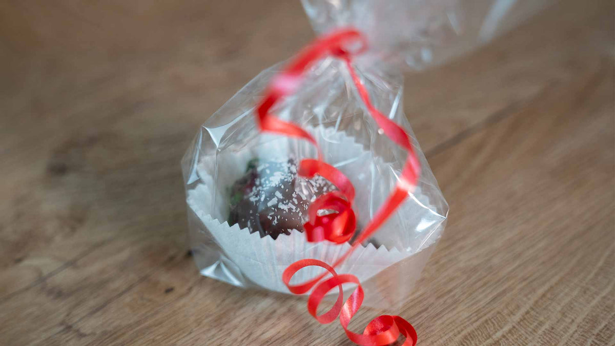 Eine mit Schokolade überzogene Leckerei in einem weißen Pappbecher ist in durchsichtiges Zellophan eingewickelt, mit einer roten Schleife zusammengebunden und auf einer Holzoberfläche platziert.