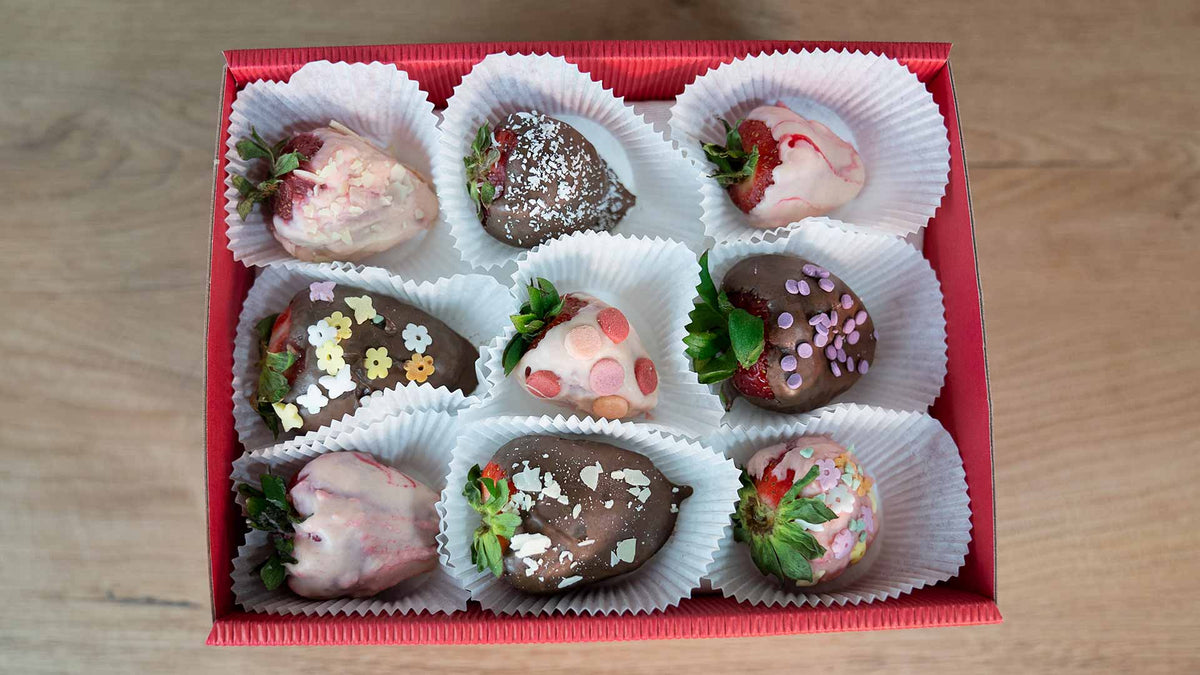 Eine rote Schachtel mit neun mit Schokolade überzogenen Erdbeeren, jede mit bunten Streuseln und Toppings verziert, angeordnet in weißen Pappbechern auf einer Holzoberfläche.