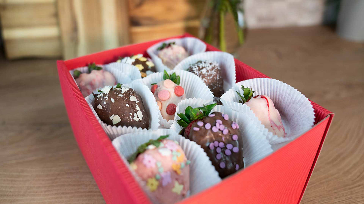 Eine rote Schachtel gefüllt mit schokoladenüberzogenen Erdbeeren, jede mit bunten Streuseln, Sternen und Herzen verziert, angeordnet in weißen Pappbechern auf einer Holzoberfläche.